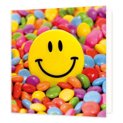 Smarties - Formaat: Standaard - Vorm: Vierkant - Blanco kaart - Op deze wenskaart staat een illustratie van een grote, gele smileyknop centraal afgebeeld, omringd door een kleurrijke zee van kleine, gekleurde snoepjes in diverse tinten zoals rood, blauw, groen en paars. De smiley heeft twee zwarte ovale ogen en een brede, zwarte glimlach. Er is geen tekst op de illustratie zichtbaar. - Snelwenskaart