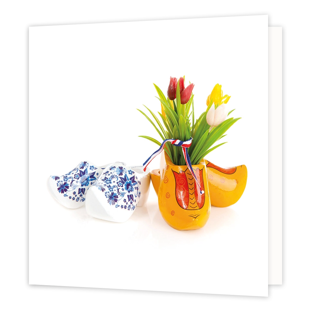 Klompen - Formaat: Standaard - Vorm: Vierkant - Blanco kaart - Op deze wenskaart staat een illustratie van twee paar houten klompen tegen een witte achtergrond. Links ligt een paar witte klompen versierd met blauwe bloemmotieven, en rechts staan gele klompen, waarvan de voorste dient als vaas gevuld met gekleurde tulpen en een rood-wit-blauw strikje. Er is geen tekst op de illustratie zichtbaar. - Snelwenskaart