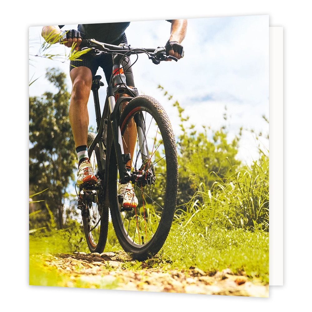Mountainbike - Formaat: Standaard - Vorm: Vierkant - Blanco kaart - Op deze wenskaart staat een illustratie van een mountainbiker die over een hobbelig bospad rijdt, van dichtbij gefotografeerd vanuit een laag standpunt. De fietser draagt sportkleding, handschoenen en opvallende schoenen, omringd door groene begroeiing en bomen tegen een lichte lucht. Er is geen tekst op de illustratie zichtbaar. - Snelwenskaart