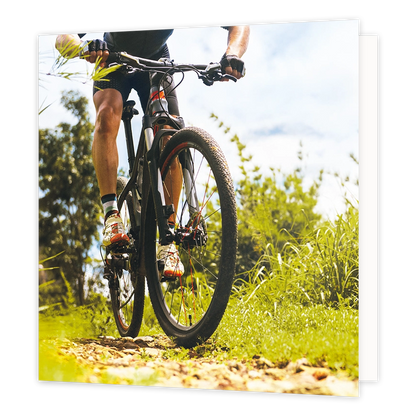 Mountainbike - Formaat: Standaard - Vorm: Vierkant - Blanco kaart - Op deze wenskaart staat een illustratie van een mountainbiker die over een hobbelig bospad rijdt, van dichtbij gefotografeerd vanuit een laag standpunt. De fietser draagt sportkleding, handschoenen en opvallende schoenen, omringd door groene begroeiing en bomen tegen een lichte lucht. Er is geen tekst op de illustratie zichtbaar. - Snelwenskaart