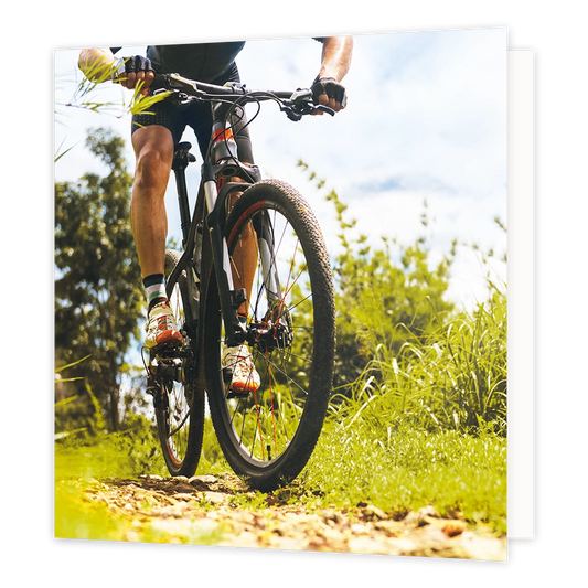 Mountainbike - Formaat: Standaard - Vorm: Vierkant - Blanco kaart - Op deze wenskaart staat een illustratie van een mountainbiker die over een hobbelig bospad rijdt, van dichtbij gefotografeerd vanuit een laag standpunt. De fietser draagt sportkleding, handschoenen en opvallende schoenen, omringd door groene begroeiing en bomen tegen een lichte lucht. Er is geen tekst op de illustratie zichtbaar. - Snelwenskaart