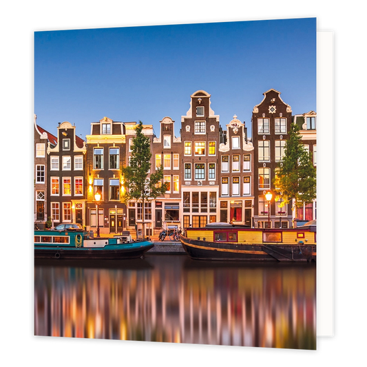 Amsterdamse grachten - Formaat: Standaard - Vorm: Vierkant - Blanco kaart - Op deze wenskaart staat een illustratie van historische Amsterdamse grachtenpanden tijdens de schemering, met sfeervol verlichte ramen en straatlantaarns. Langs de kade en in het water liggen woonboten, en de lichten van de stad weerspiegelen prachtig in het water. De hemel erboven is helderblauw en er is geen tekst op de illustratie zichtbaar. - Snelwenskaart