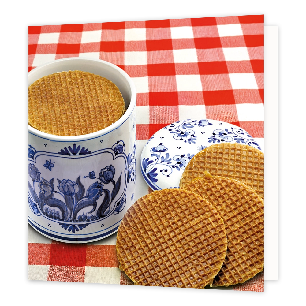 Stroopwafels - Formaat: Standaard - Vorm: Vierkant - Blanco kaart - Op deze wenskaart staat een illustratie van een traditionele blauw-witte Delfts blauwe voorraadpot gevuld met stroopwafels, geplaatst op een rood-wit geruite tafel. Naast de pot ligt het bijbehorende deksel met een bloemmotief en zijn er nog drie stroopwafels voor de pot neergelegd. Er is geen tekst op de illustratie zichtbaar. - Snelwenskaart