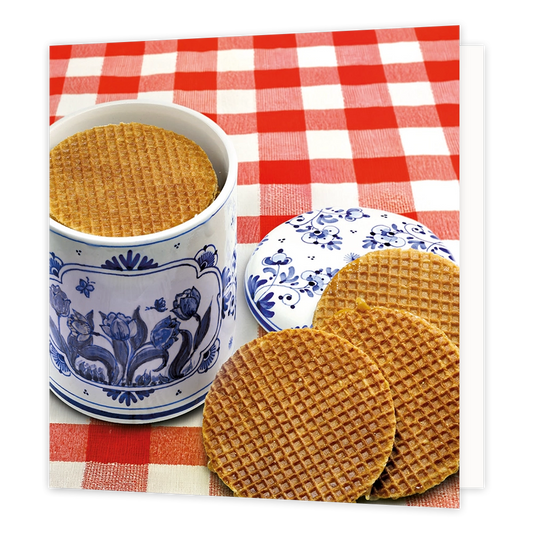 Stroopwafels - Formaat: Standaard - Vorm: Vierkant - Blanco kaart - Op deze wenskaart staat een illustratie van een traditionele blauw-witte Delfts blauwe voorraadpot gevuld met stroopwafels, geplaatst op een rood-wit geruite tafel. Naast de pot ligt het bijbehorende deksel met een bloemmotief en zijn er nog drie stroopwafels voor de pot neergelegd. Er is geen tekst op de illustratie zichtbaar. - Snelwenskaart