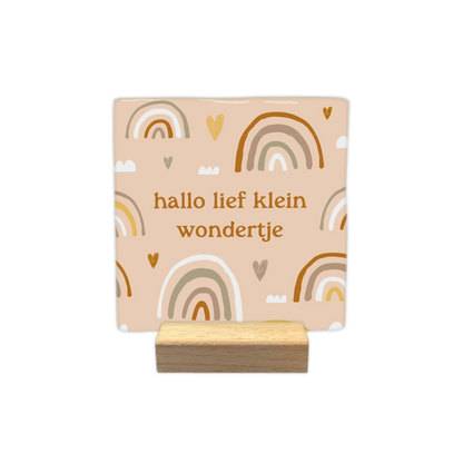 Hallo lief klein wondertje - Decoratie tegel - Formaat: - Op deze decoratietegel staat in het midden de Nederlandse tekst "hallo lief klein wondertje". De tegel zelf is beige en vierkant, en staat in een houten standaard. De tekst is verdeeld over twee regels en geschreven in een bruin lettertype in kleine letters. De achtergrond is versierd met een patroon van getekende regenbogen in aardetinten (zoals bruin, okergeel, beige en wit), gecombineerd met kleine hartjes en wolkjes.&nbsp; - Snelwenskaart