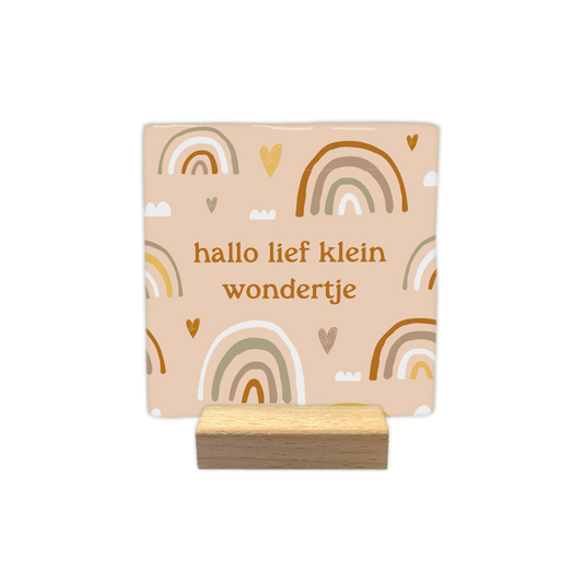 Hallo lief klein wondertje - Decoratie tegel - Formaat: - Op deze decoratietegel staat in het midden de Nederlandse tekst "hallo lief klein wondertje". De tegel zelf is beige en vierkant, en staat in een houten standaard. De tekst is verdeeld over twee regels en geschreven in een bruin lettertype in kleine letters. De achtergrond is versierd met een patroon van getekende regenbogen in aardetinten (zoals bruin, okergeel, beige en wit), gecombineerd met kleine hartjes en wolkjes.&nbsp; - Snelwenskaart