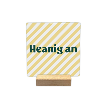Heanig an - Decoratie tegel - Formaat: - Op deze decoratie tegel staat de tekst "Heanig an" in donkergroene letters centraal in beeld. De achtergrond bestaat uit een patroon van diagonaal lopende, lichtgele en witte strepen. Het ontwerp oogt hierdoor rustig en gestreept. - Snelwenskaart