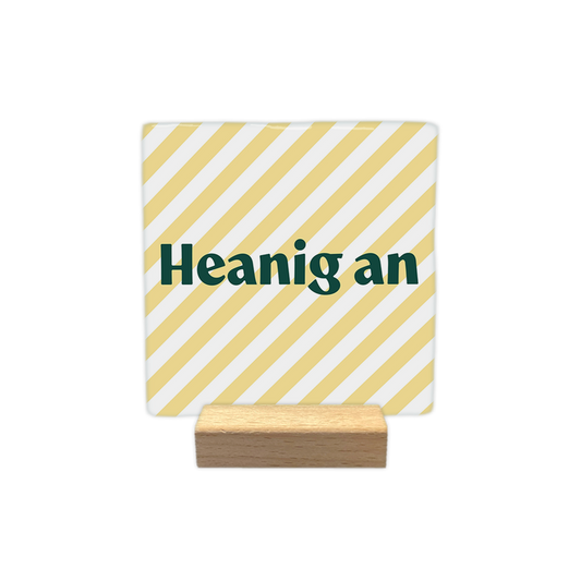 Heanig an - Decoratie tegel - Formaat: - Op deze decoratie tegel staat de tekst "Heanig an" in donkergroene letters centraal in beeld. De achtergrond bestaat uit een patroon van diagonaal lopende, lichtgele en witte strepen. Het ontwerp oogt hierdoor rustig en gestreept. - Snelwenskaart