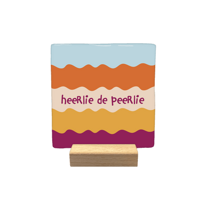 Heerlie de peerlie - Decoratie tegel - Formaat: - Op deze decoratietegel staat de tekst "heeRlie de peeRlie" in donkerpaarse, speelse letters centraal afgebeeld. De achtergrond bestaat uit horizontale, golvende banen in diverse kleuren, waaronder lichtblauw, oranje, beige, geel en donkerpaars. - Snelwenskaart