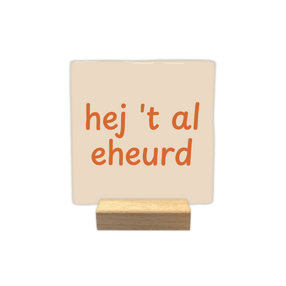 Hej 't al eheurd - Decoratie tegel - Formaat: - Op deze decoratie tegel staat de tekst "hej 't al eheurd" in oranje letters verdeeld over twee regels. Het ontwerp maakt gebruik van een speels lettertype en is geplaatst op een lichtbeige achtergrond. - Snelwenskaart