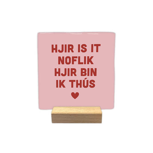 Hjir is it noflik hjir bin ik thús - Decoratie tegel - Formaat: - Op deze decoratie tegel staat de tekst "hjir is it noflik hjir bin ik thús" in rode letters centraal afgebeeld. Onder de tekst is een klein rood hartje geplaatst. De achtergrond van de tegel is lichtroze van kleur. - Snelwenskaart