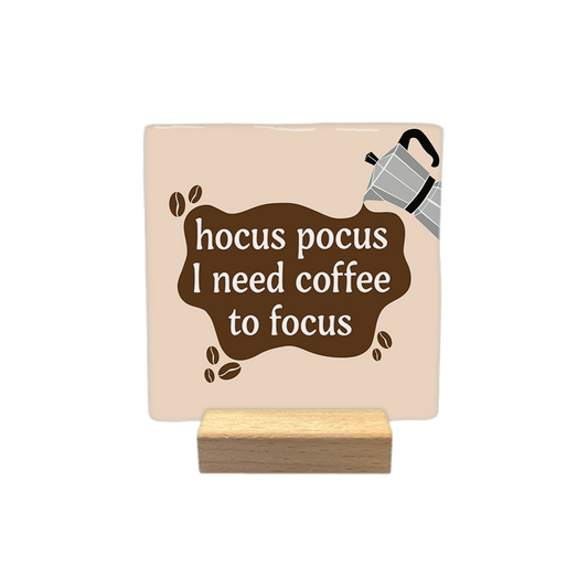 Hocus pocus I need coffee to focus - Decoratie tegel - Formaat: - Op deze decoratietegel staat in een bruine, onregelmatige vorm de tekst "hocus pocus I need coffee to focus" in witte letters. Rechtsboven deze vorm wordt een grijze percolator afgebeeld waaruit de bruine kleur lijkt te vloeien. Het ontwerp is geplaatst op een beige, vierkante achtergrond en bevat enkele kleine koffieboontjes als decoratie. - Snelwenskaart