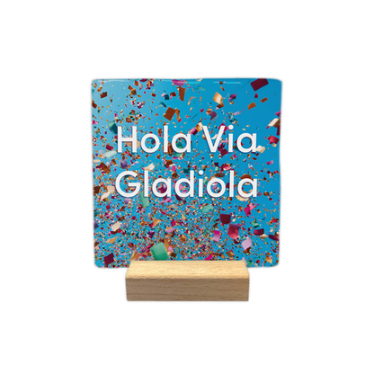 Hola via Gladiola - Decoratie tegel - Formaat: - Op deze decoratie tegel staat de tekst "Hola Via Gladiola" in witte letters centraal in beeld. De achtergrond is felblauw en gevuld met kleurrijke, rondvliegende confetti. Het ontwerp geeft een feestelijke indruk door de verspreiding van de snippers over het gehele vlak. - Snelwenskaart