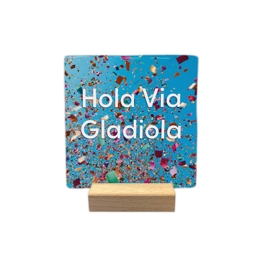 Hola via Gladiola - Decoratie tegel - Formaat: - Op deze decoratie tegel staat de tekst "Hola Via Gladiola" in witte letters centraal in beeld. De achtergrond is felblauw en gevuld met kleurrijke, rondvliegende confetti. Het ontwerp geeft een feestelijke indruk door de verspreiding van de snippers over het gehele vlak. - Snelwenskaart