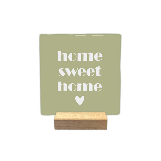 Home sweet home - Decoratietegel – 10 x 10 cm - Op deze decoratietegel staat in het midden de Engelse tekst "home sweet home". De tegel zelf is olijfgroen en vierkant, en staat in een houten standaard. De tekst is verdeeld over drie regels en geschreven in een strak, wit lettertype in kleine letters, met daaronder een klein, wit hartje gecentreerd in het midden.