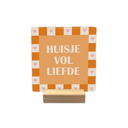 Huisje vol liefde - Decoratie tegel - Formaat: - Op deze decoratie tegel staat centraal de tekst "HUISJE VOL LIEFDE" in witte letters op een oranje achtergrond. De illustratie is omrand door een schaakbordpatroon van afwisselend oranje en lichtbeige vierkanten. In elk van de lichtbeige vierkanten van de rand is een klein roze hartje afgebeeld. - Snelwenskaart