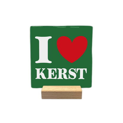 I love kerst - Decoratie tegel - Formaat: - Op deze decoratie tegel staat een groene achtergrond met witte en rode elementen. Links staat een witte letter i en rechts daarvan is een rood hart te zien. Onder dit hart en de letter staat in witte letters het woord kerst. De tegel staat op een kleine houten houder. - Snelwenskaart