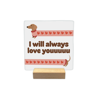 I will always love youuuuu - Decoratie tegel - Formaat: - Op deze decoratie tegel staat een illustratie van een extreem langgerekte teckel die over de hele breedte van de kaart uitgerekt lijkt. Het lichaam van de hond is gevuld met een patroon van kleine rode hartjes op een lichtroze achtergrond. In het midden van de afbeelding staat in dikke, bruine letters de tekst "I will always love youuuuu". - Snelwenskaart