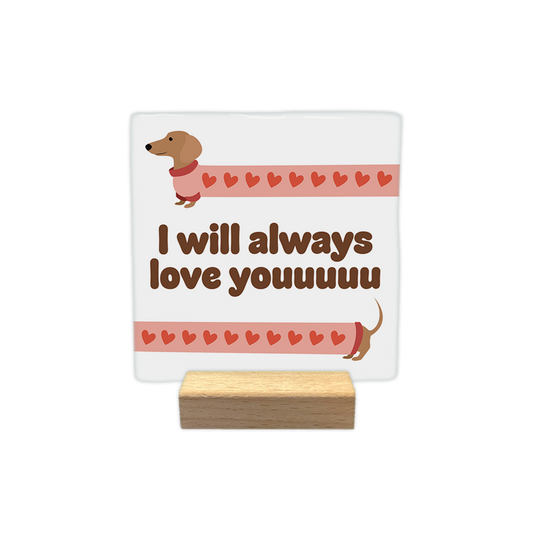 I will always love youuuuu - Decoratie tegel - Formaat: - Op deze decoratie tegel staat een illustratie van een extreem langgerekte teckel die over de hele breedte van de kaart uitgerekt lijkt. Het lichaam van de hond is gevuld met een patroon van kleine rode hartjes op een lichtroze achtergrond. In het midden van de afbeelding staat in dikke, bruine letters de tekst "I will always love youuuuu". - Snelwenskaart