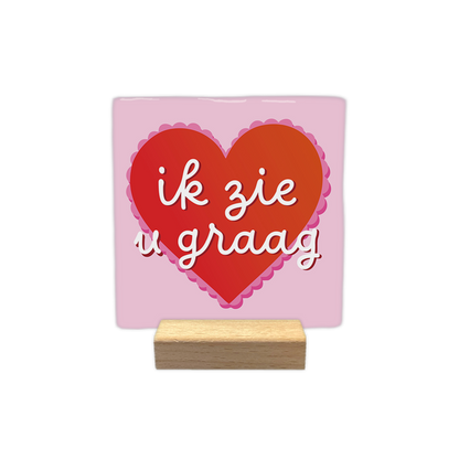 Ik zie u graag - Decoratie tegel - Formaat: - Op deze decoratie tegel staat de tekst "ik zie u graag" in witte, handgeschreven letters afgebeeld. De tekst is geplaatst in een rood hart dat een roze, geschulpte rand heeft. De achtergrond van de tegel is lichtroze van kleur. - Snelwenskaart