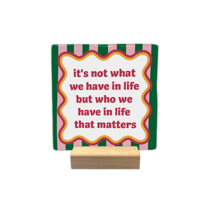 It's not what we have in life - Decoratie tegel - Formaat: - Op deze decoratie tegel staat centraal de tekst "it's not what we have in life but who we have in life that matters" in roze letters binnen een wit kader met een golvende, meerlaagse rand in de kleuren oranje, roze en donkerrood. De achtergrond van het ontwerp bestaat uit verticale strepen in afwisselende tinten donkergroen en roze. - Snelwenskaart