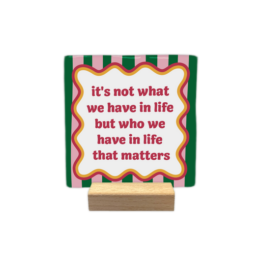 It's not what we have in life - Decoratie tegel - Formaat: - Op deze decoratie tegel staat centraal de tekst "it's not what we have in life but who we have in life that matters" in roze letters binnen een wit kader met een golvende, meerlaagse rand in de kleuren oranje, roze en donkerrood. De achtergrond van het ontwerp bestaat uit verticale strepen in afwisselende tinten donkergroen en roze. - Snelwenskaart