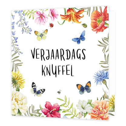 Verjaardagsknuffel - Formaat: Standaard - Vorm: Vierkant - Verjaardagskaart - Op deze wenskaart staat in het midden in zwarte, handgeschreven letters de tekst "VERJAARDAGS KNUFFEL". De tekst wordt omringd door een kleurrijke rand van diverse bloemen, waaronder roze, oranje, gele en blauwe varianten, afgewisseld met groen blad. Tussen de bloemen vliegen verschillende vlinders, een lieveheersbeestje en een bijtje afgebeeld. De illustratie heeft een witte achtergrond en is vormgegeven in een botanische aquare