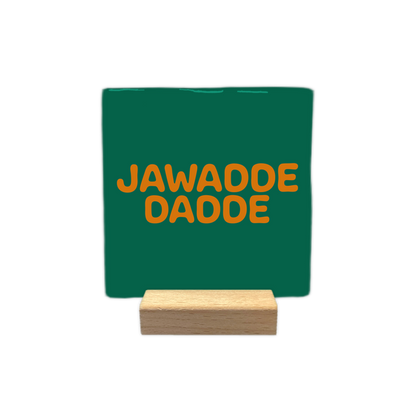 Jawadde dadde - Decoratie tegel - Formaat: - Op deze decoratie tegel staat de tekst "jawadde dadde" in twee regels afgebeeld. De tekst is geschreven in oranje, afgeronde letters. De achtergrond van de tegel is donkergroen. - Snelwenskaart