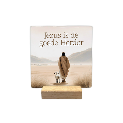 Jezus is de goede herder - Decoratie tegel - Formaat: - Op deze decoratie tegel staat bovenaan de tekst "Jezus is de goede Herder". Daaronder is een afbeelding te zien van een figuur in een gewaad die in een woestijnachtig landschap loopt met een lammetje aan zijn zijde. De omgeving bevat zand, gras en bergen op de achtergrond in zachte, beige tinten. - Snelwenskaart