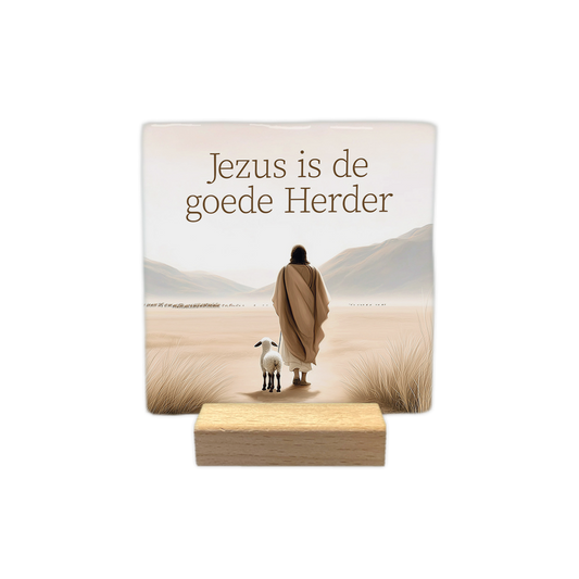 Jezus is de goede herder - Decoratie tegel - Formaat: - Op deze decoratie tegel staat bovenaan de tekst "Jezus is de goede Herder". Daaronder is een afbeelding te zien van een figuur in een gewaad die in een woestijnachtig landschap loopt met een lammetje aan zijn zijde. De omgeving bevat zand, gras en bergen op de achtergrond in zachte, beige tinten. - Snelwenskaart