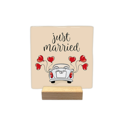 Just married - Decoratie tegel - Formaat: - Op deze decoratie tegel staat een illustratie van de achterkant van een witte trouwauto op een beige achtergrond. De auto is versierd met een strik en twee ringen op de achterklep, en er zijn vijf rode hartvormige ballonnen aan verbonden. Bovenaan de afbeelding staat in sierlijke, zwarte letters de tekst "just married" geschreven. - Snelwenskaart
