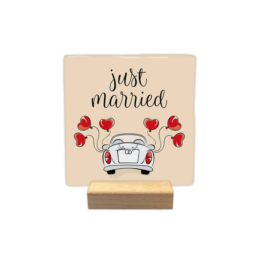 Just married - Decoratie tegel - Formaat: - Op deze decoratie tegel staat een illustratie van de achterkant van een witte trouwauto op een beige achtergrond. De auto is versierd met een strik en twee ringen op de achterklep, en er zijn vijf rode hartvormige ballonnen aan verbonden. Bovenaan de afbeelding staat in sierlijke, zwarte letters de tekst "just married" geschreven. - Snelwenskaart