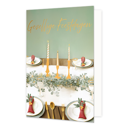 Gezellige feestdagen - Formaat: Standaard - Vorm: Staand - Kerstkaart - Op deze wenskaart staat een feestelijk gedekte tafel afgebeeld met borden, bestek, glazen en rode servetten met takjes erin. In het midden van de tafel loopt een groene slinger met drie gouden kandelaars waarin brandende kaarsen staan. De achtergrond is zachtgroen van kleur en toont de sfeer van een feestelijke maaltijd. Bovenaan de illustratie staat de gele, handgeschreven tekst "Gezellige Feestdagen". - Snelwenskaart