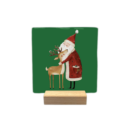 Kerstman en rendier - Decoratie tegel - Formaat: - Op deze decoratie tegel staat een groene achtergrond met een geïllustreerde kerstman die een rendier knuffelt. De kerstman draagt een rode jas en een rode muts met een witte rand en pompon. Het rendier heeft een bruine vacht en een gewei met kleine takjes. - Snelwenskaart