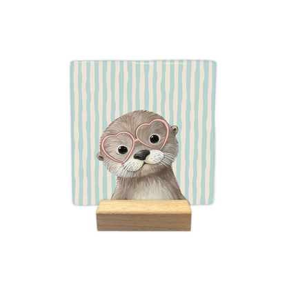 Kijkende otter - Decoratie tegel - Formaat: - Op deze decoratie tegel staat een otter afgebeeld die een roze bril met hartvormige glazen draagt. Het dier is van voren te zien en kijkt recht naar voren. De achtergrond van de tegel is versierd met verticale lijnen in de kleuren lichtblauw en beige. - Snelwenskaart