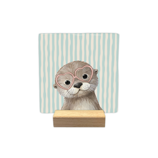 Kijkende otter - Decoratie tegel - Formaat: - Op deze decoratie tegel staat een otter afgebeeld die een roze bril met hartvormige glazen draagt. Het dier is van voren te zien en kijkt recht naar voren. De achtergrond van de tegel is versierd met verticale lijnen in de kleuren lichtblauw en beige. - Snelwenskaart