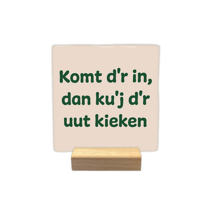 Komt d'r in, dan ku'j d'r uut kieken - Decoratie tegel - Formaat: - Op deze decoratie tegel staat de tekst "Komt d'r in, dan ku'j d'r uut kieken" verdeeld over drie regels. De letters zijn donkergroen en in een speels lettertype weergegeven. De achtergrond heeft een lichtbeige kleur. - Snelwenskaart