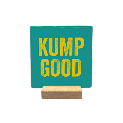 Kump Good - Decoratie tegel - Formaat: - Op deze decoratie tegel staat de tekst "kump good" in gele letters. De tekst is verdeeld over twee regels en geplaatst op een blauwgroene achtergrond. De letters hebben een textuur die eruitziet alsof ze gestempeld of verweerd zijn. - Snelwenskaart