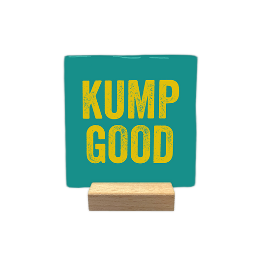 Kump Good - Decoratie tegel - Formaat: - Op deze decoratie tegel staat de tekst "kump good" in gele letters. De tekst is verdeeld over twee regels en geplaatst op een blauwgroene achtergrond. De letters hebben een textuur die eruitziet alsof ze gestempeld of verweerd zijn. - Snelwenskaart