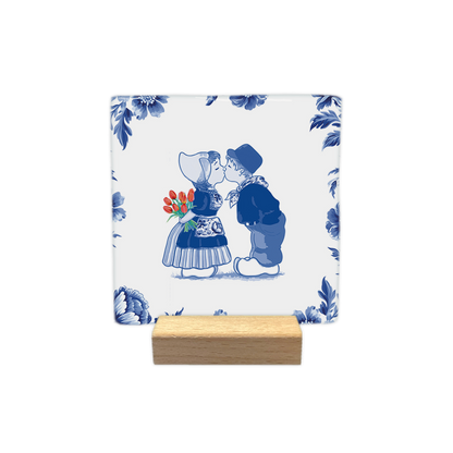 Kussend paar - Decoratie tegel - Formaat: - Op deze decoratie tegel staat een illustratie van een kussend jongetje en meisje in traditionele Nederlandse klederdracht, omringd door een rand van Delfts blauwe bloemen op een witte achtergrond. Het meisje draagt een kanten mutsje en houdt een boeket rode tulpen achter haar rug vast. De jongen draagt een pet en beide kinderen dragen klompen. De gehele afbeelding is uitgevoerd in een klassieke blauw-witte kleurstelling, met uitzondering van de rode tulpen. - Sne
