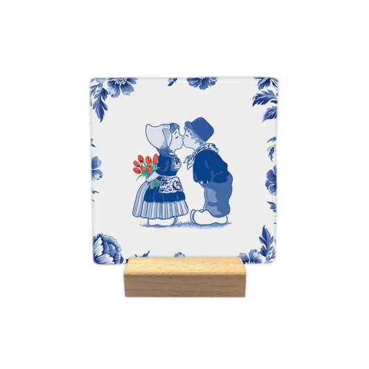 Kussend paar - Decoratie tegel - Formaat: - Op deze decoratie tegel staat een illustratie van een kussend jongetje en meisje in traditionele Nederlandse klederdracht, omringd door een rand van Delfts blauwe bloemen op een witte achtergrond. Het meisje draagt een kanten mutsje en houdt een boeket rode tulpen achter haar rug vast. De jongen draagt een pet en beide kinderen dragen klompen. De gehele afbeelding is uitgevoerd in een klassieke blauw-witte kleurstelling, met uitzondering van de rode tulpen. - Sne