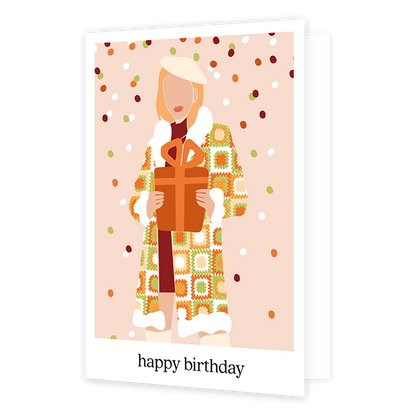 Happy birthday - Formaat: Normaal - Vorm: Staand - Verjaardagskaart - Op deze wenskaart staat een vrouw met een lange jas aan en een groot cadeau in haar handen. De achtergrond is zacht roze met groen, rood, oranje en witte sneeuwval. De afbeelding staat in een witkader en onderin staat de tekst 'happy birthday'. - Snelwenskaart