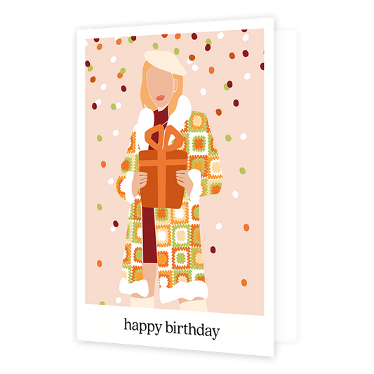 Happy birthday - Formaat: Normaal - Vorm: Staand - Verjaardagskaart - Op deze wenskaart staat een vrouw met een lange jas aan en een groot cadeau in haar handen. De achtergrond is zacht roze met groen, rood, oranje en witte sneeuwval. De afbeelding staat in een witkader en onderin staat de tekst 'happy birthday'. - Snelwenskaart