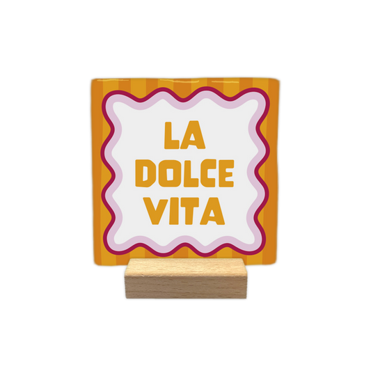 La dolce vita - Decoratie tegel - Formaat: - Op deze decoratie tegel staat centraal de tekst "LA DOLCE VITA" in gele, blokkerige letters binnen een wit kader met een golvende rand. Het kader is omlijnd met roze en donkerrode lijnen. De achtergrond van het ontwerp bestaat uit verticale strepen in afwisselende tinten oranje. - Snelwenskaart