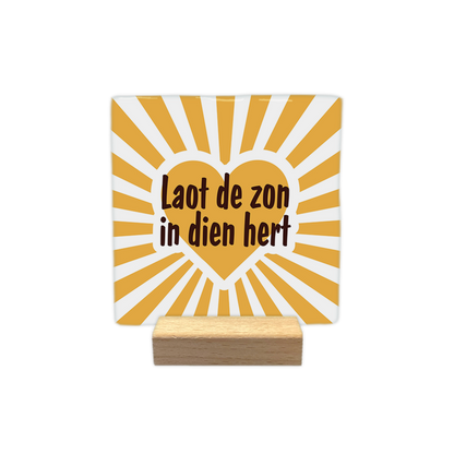 Laot de zon in dien - Decoratie tegel - Formaat: - Op deze decoratie tegel staat centraal een geel hart met daarin de tekst "Laot de zon in dien hert" in donkerbruine letters. De achtergrond bestaat uit een stralend patroon van gele en witte lijnen die vanuit het midden naar buiten lopen. Het ontwerp heeft een opgewekte uitstraling door het gebruik van het hart en de zonnestralen. - Snelwenskaart