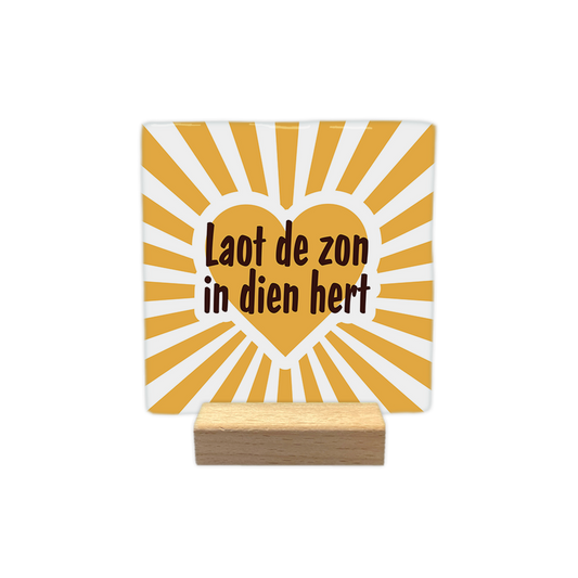 Laot de zon in dien - Decoratie tegel - Formaat: - Op deze decoratie tegel staat centraal een geel hart met daarin de tekst "Laot de zon in dien hert" in donkerbruine letters. De achtergrond bestaat uit een stralend patroon van gele en witte lijnen die vanuit het midden naar buiten lopen. Het ontwerp heeft een opgewekte uitstraling door het gebruik van het hart en de zonnestralen. - Snelwenskaart