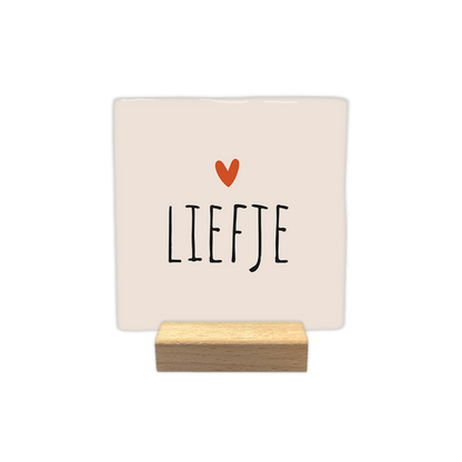 Liefje - Decoratie tegel - Formaat: - Op deze decoratie tegel staat de tekst "liefje" in zwarte letters afgebeeld. Boven de tekst bevindt zich een klein oranje hartje. De achtergrond van de tegel is lichtbeige van kleur. - Snelwenskaart