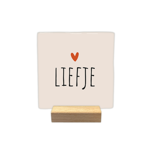 Liefje - Decoratie tegel - Formaat: - Op deze decoratie tegel staat de tekst "liefje" in zwarte letters afgebeeld. Boven de tekst bevindt zich een klein oranje hartje. De achtergrond van de tegel is lichtbeige van kleur. - Snelwenskaart
