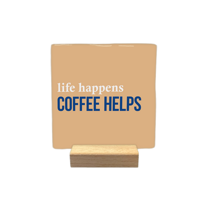 Life happens coffee helps - Decoratie tegel - Formaat: - Op deze decoratietegel staat de tekst "life happens" in witte letters bovenaan, met daaronder in grotere, donkerblauwe hoofdletters "COFFEE HELPS". Het ontwerp is gecentreerd op een lichtbruine achtergrond. - Snelwenskaart