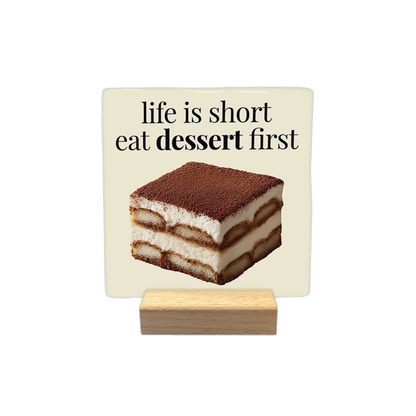 Life is short eat dessert first - Decoratie tegel - Formaat: - Op deze decoratietegel staat de tekst "life is short eat dessert first" in zwarte letters met een schreeflettertype. Daaronder is een realistische afbeelding van een vierkant stuk tiramisu geplaatst. Het ontwerp is gecentreerd op een crèmekleurige achtergrond. - Snelwenskaart